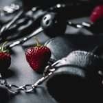 Close-up of a strawberry amidst chains and leather on a dark surface, artistic composition.